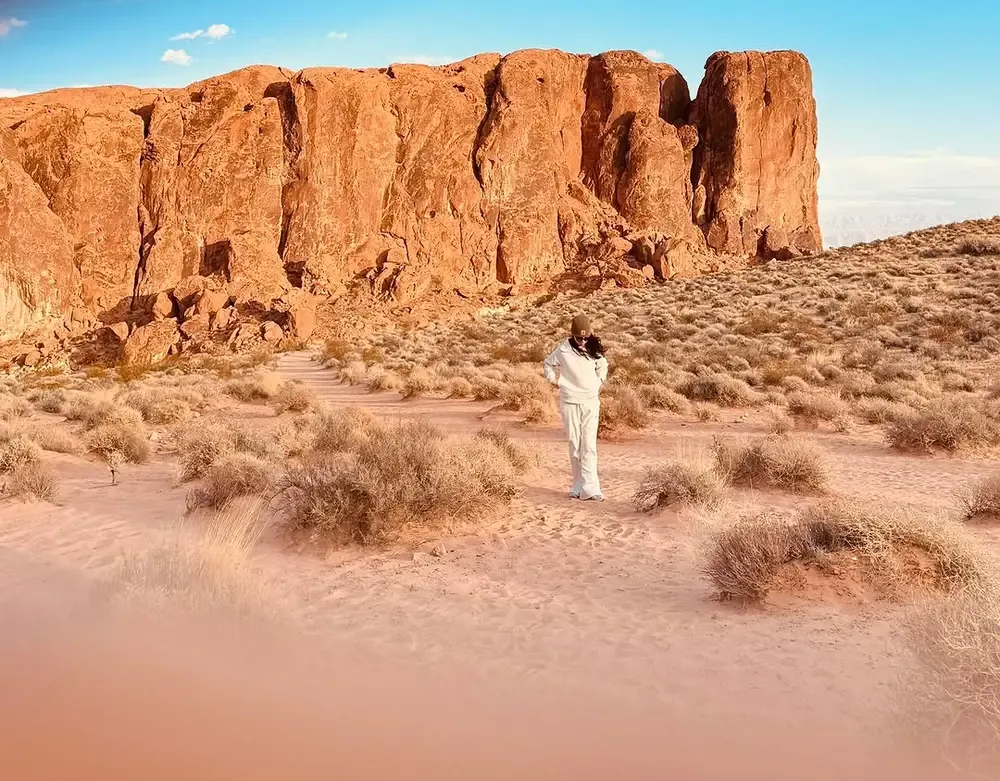 Visitor walking a sandy trail beneath a tall red sandstone wall in Valley of Fire State Park, Nevada
